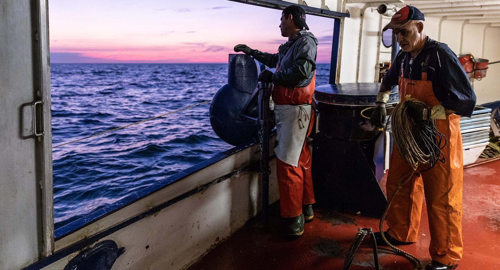 Two fishermen working on deck at dawn, with the sea and a pink sky through the open side of the boat