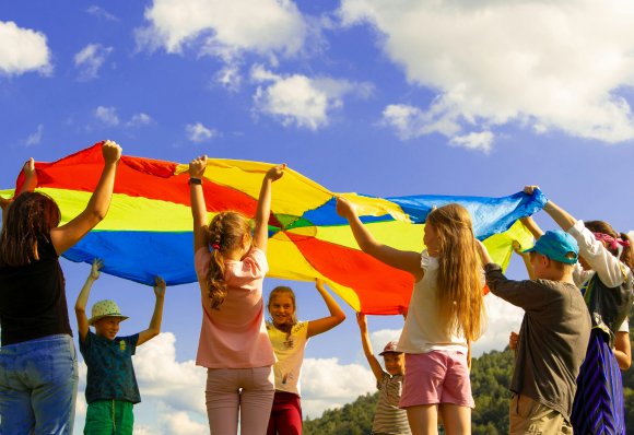 Children holding a rainbow play parachute overhead in a sunny field